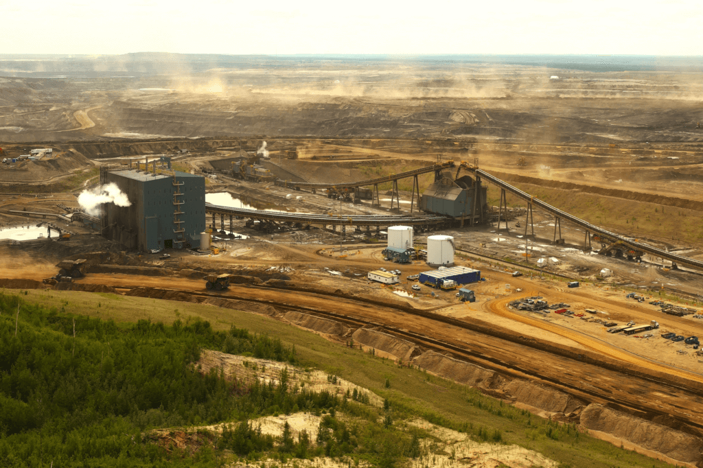 A large-scale open-pit mining operation with extensive excavated terrain in the background. Prominent in the mid-ground are several industrial structures, including a large processing facility with visible steam or smoke emission, conveyor systems, and storage tanks. Heavy machinery like excavators and dump trucks are scattered across the site. The foreground features a green strip of forested land transitioning into dirt roads and mining pathways. The atmosphere appears hazy, possibly from dust or industrial emissions.