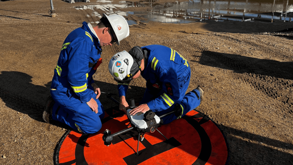 Two workers wearing blue safety coveralls, helmets, and reflective stripes are kneeling on the ground, preparing a drone for operation. The drone is positioned on a red landing pad with a black “H” symbol. One worker adjusts the drone, while the other observes closely. The setting is an outdoor industrial site with a dirt surface, tire tracks, and water puddles reflecting nearby structures. The workers appear focused and are equipped with safety gear.



