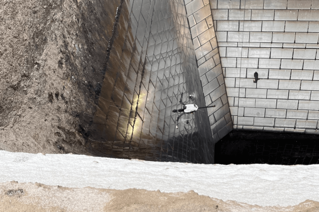  drone is seen conducting an inspection in a steep, enclosed ore bin with walls made of metallic, grid-patterned plates. The drone hovers close to the reflective metal surfaces, which appear slightly dirty or weathered. The walls of the structure curve sharply, creating a deep, narrow space. The upper portion shows an uneven, textured area of soil or debris adjacent to the metallic surfaces.